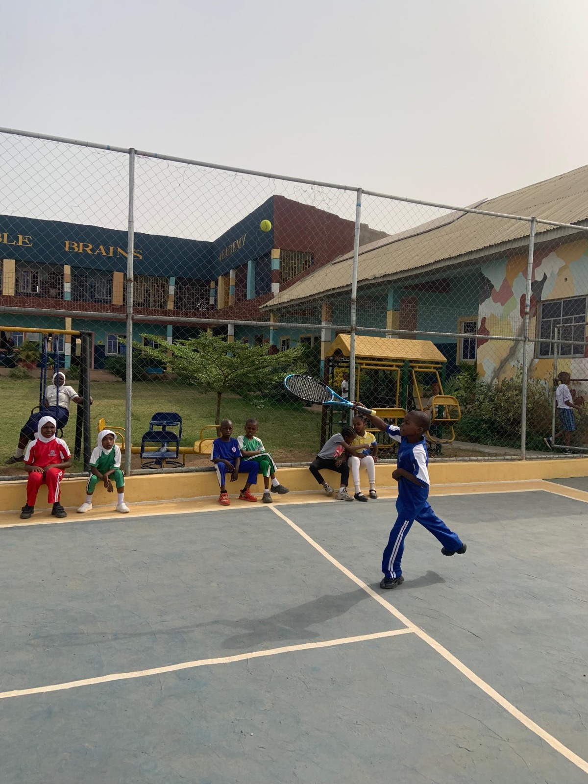 Noble Brains Academy pupil playing tennis on sports court with school building in background